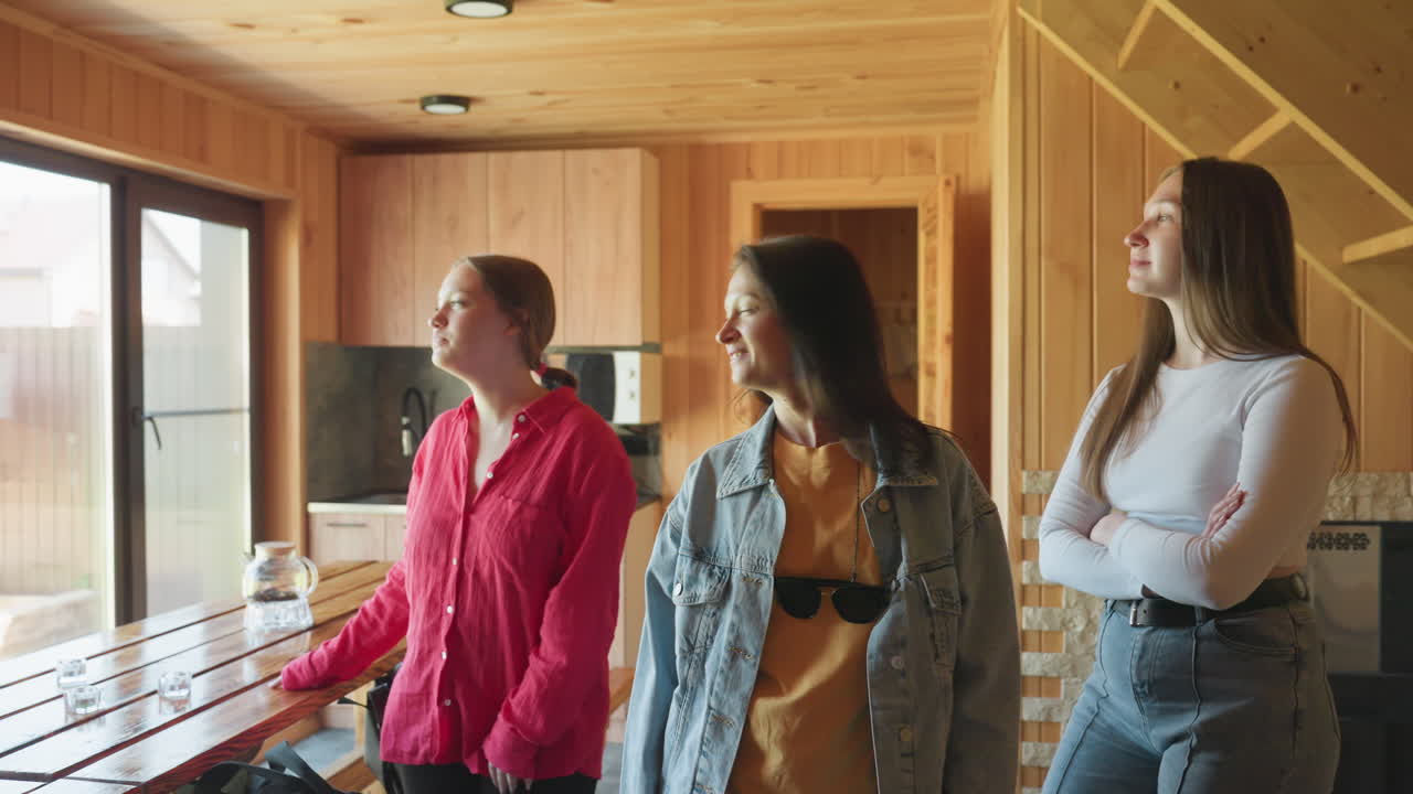 three young women stand near wooden dining table inside cabin kitchen, casually observing outdoors through window, appearing bored but smiling lightly