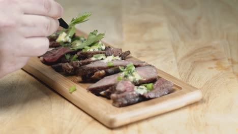 Slow-Motion-Slider-Shot-of-Medium-Rare-Sliced-Steak-on-a-Rectangular-wooden-Chopping-Board-With-Food-Stylist-Hands-Adding-Garnish