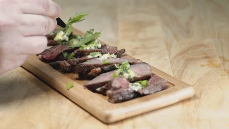 toma de control deslizante en cámara lenta de filete medio raro en rodajas en una tabla de cortar de madera rectangular con manos de estilista de alimentos agregando guarnición