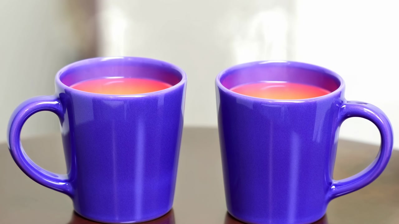 Two tall glossy purple ceramic mugs sit side by side on reflective tabletop filled with warm orange liquid while visible steam rises above each rim against soft