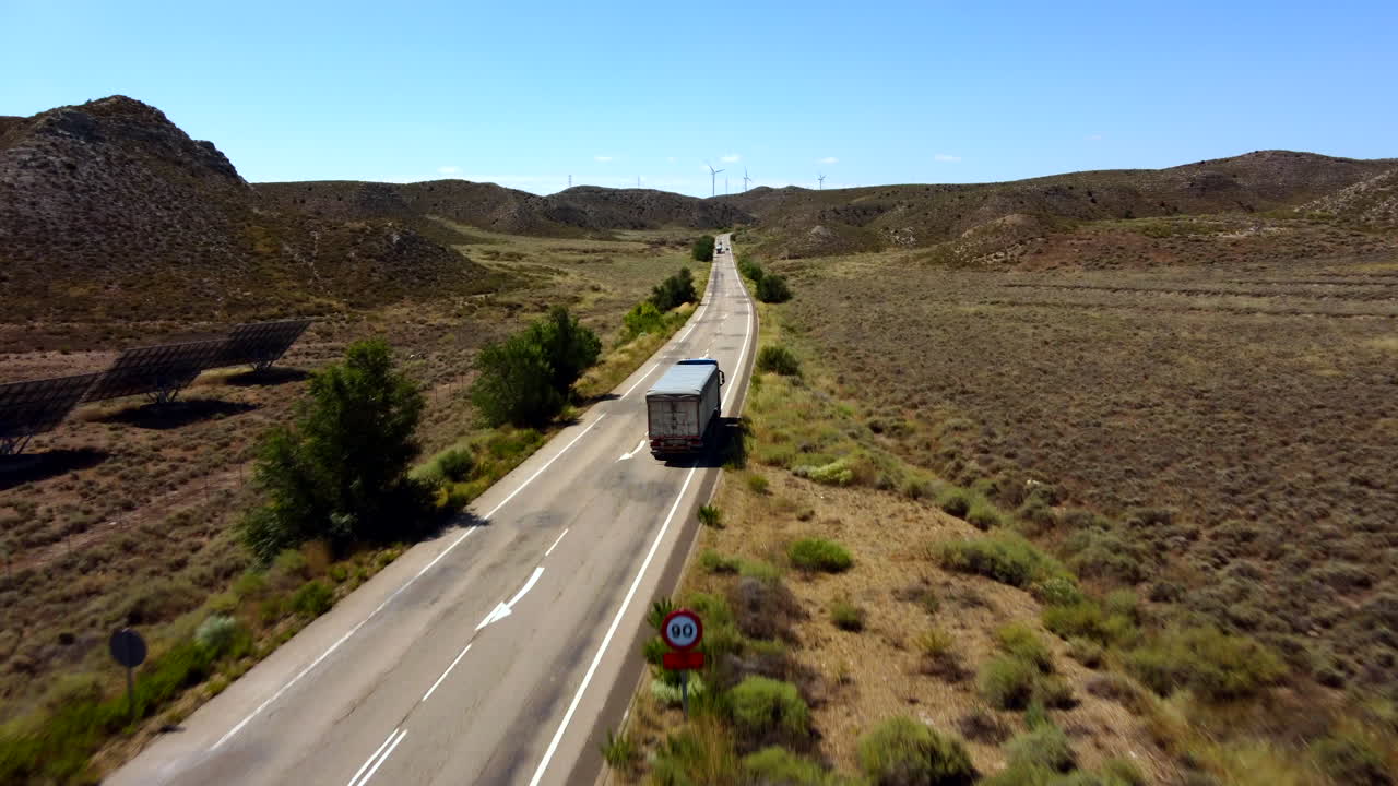 vista aerea di un'auto in viaggio su strada che sorpassa camion che percorrono un luogo deserto, con mulini a vento sullo sfondo