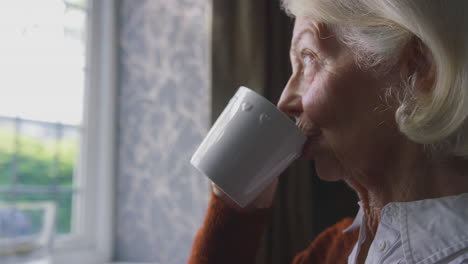 Senior-Woman-With-Hot-Drink-Trying-To-Keep-Warm-By-Radiator-At-Home-In-Cost-Of-Living-Energy-Crisis