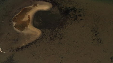 a top-down aerial view over a salt marsh, it is low tide - you can see a sandbar in the water. the camera rotates counterclockwise and pulls away from the sand mound in the dark green waters.