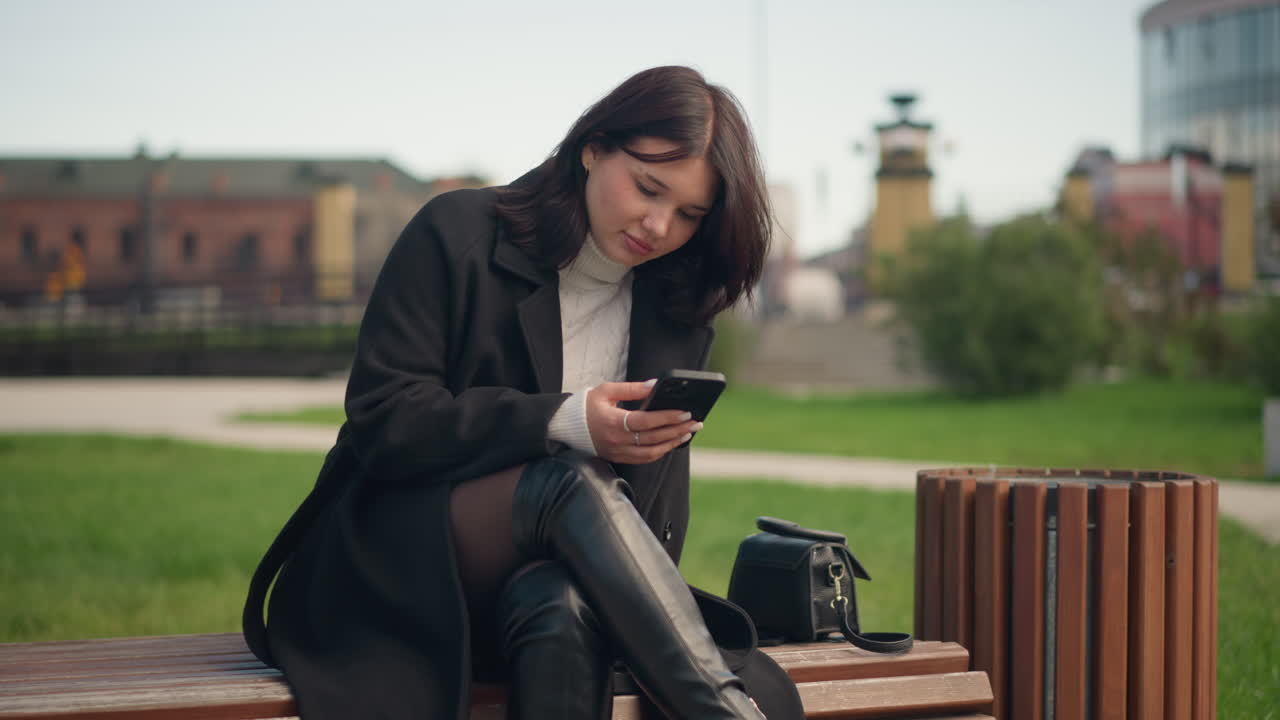 검은색 코트와 색 터틀을 입은 어린 소녀가 에서 바람 가방에 날아다니는 머리카락으로 드폰에 초점을 맞추고, 배경에 주거 건물과 함께 야외에 앉아 있습니다.