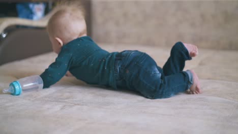 funny boy crawls with bottle on large beige bed in room