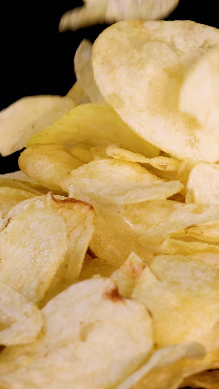 Golden potato chips drop onto a pile against black background, captured in crisp studio lighting