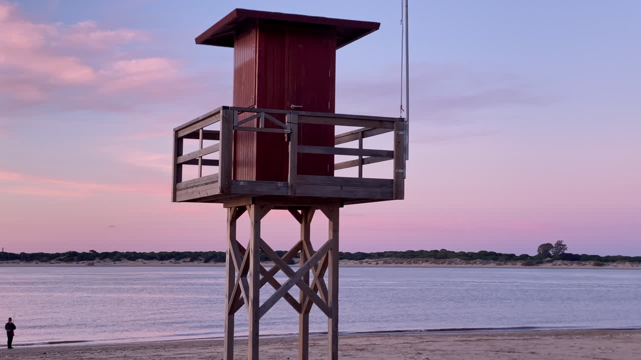 Livreddertårn på en sandstrand ved solnedgang, der viser en gradienthimmel med en flodmunding og både i baggrunden