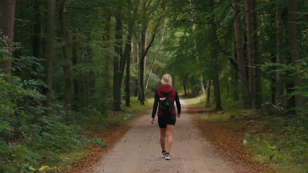 Vista traseira de uma mulher caucasiana caminhando por um caminho na floresta no verão, cercada por belas árvores verdes densas e folhagem exuberante