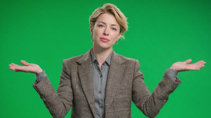 Una donna con un elegante blazer grigio gesticola con entrambe le mani, mostrando sicurezza e compostezza su uno sfondo verde vivace, suscitando curiosità in un ambiente dinamico