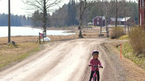 Handheld,-Zeitlupenaufnahme-Eines-Kleinen-Mädchens,-Das-Auf-Einer-Schotterstraße-Auf-Der-Finnischen-Landschaft-An-Einem-Sonnigen-Frühlingstag-In-Vaasa,-Ostbottnien,-Finnland,-Rad-Fährt-1