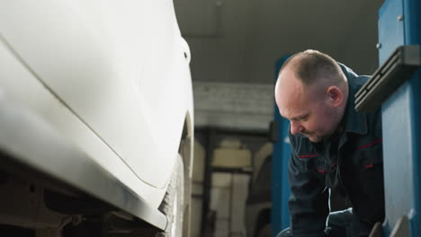 ingeniero automotriz en uniforme azul y guantes se inclina para colocar un elevador de automóvil bajo un vehículo blanco en un taller con un poste de elevación azul visible en el fondo, preparándose para el mantenimiento