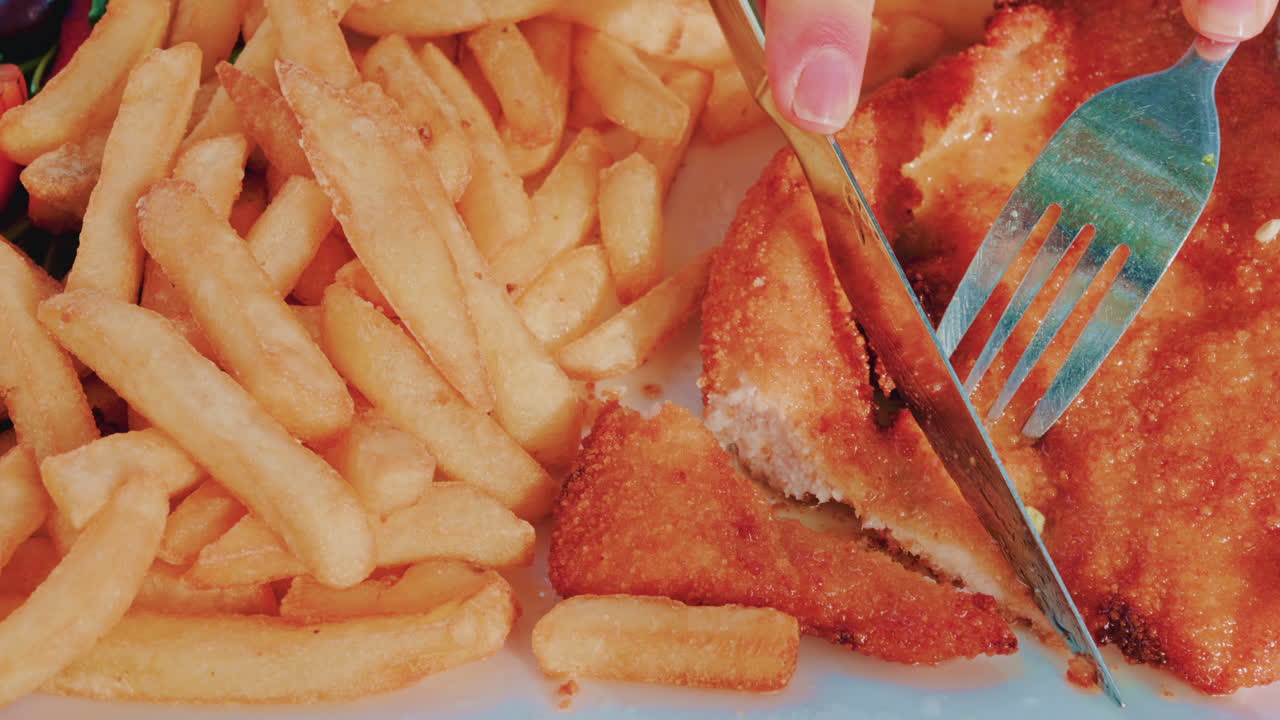 Crispy breaded fillet served with golden French fries on a blue plate as a hands cut the meat with a fork and knife
