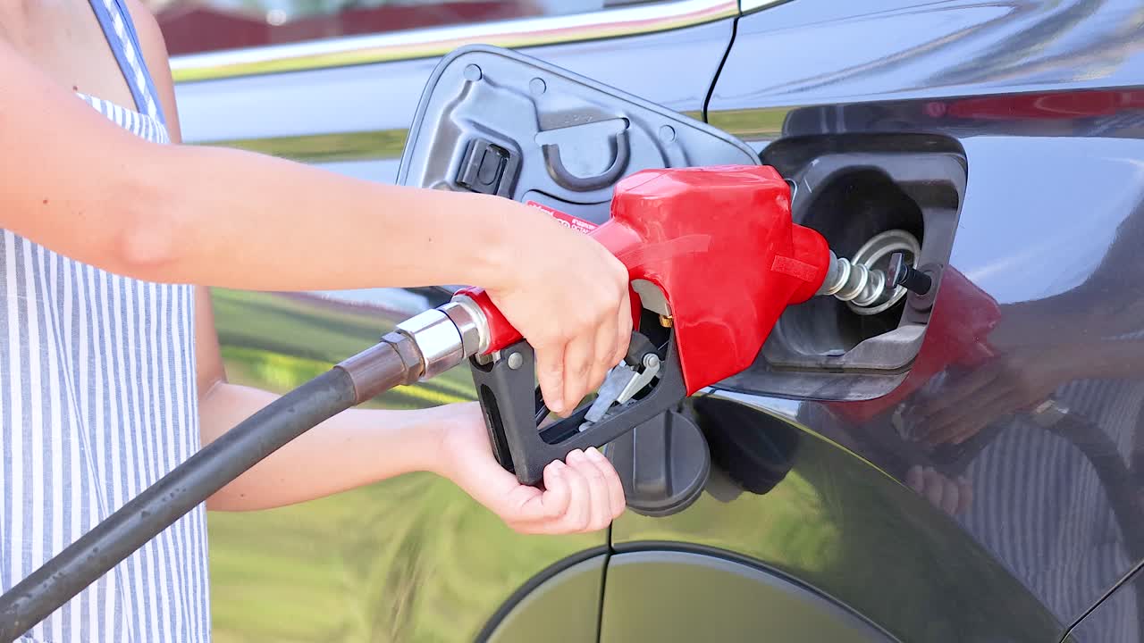 A person refuels a car at a coin-operated dispenser in bright daylight, highlighting the vibrant red nozzle