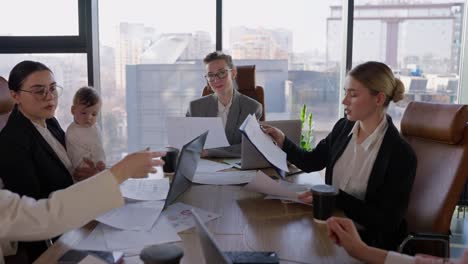 portrait of a confident middle-aged blonde business woman wearing glasses in a gray suit while she sits at an office desk during a businesswoman meeting in a modern office with large windows