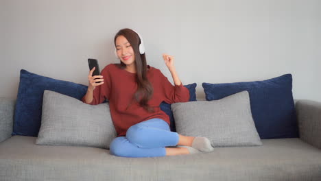 Young-happy-asian-woman-enjoys-in-music-from-smartphone-with-wireless-headphones-on-couch-in-living-room,-full-frame-slow-motion