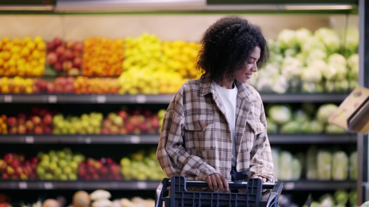 Cheerful woman walks through supermarket with cart, taking a pack of potatoes