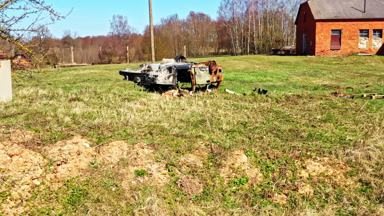 Wide shot of grassy rural field with burned car wreckage and old brick house in background