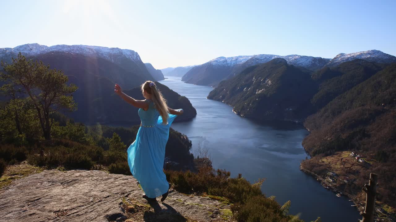Young attractive woman spins, dances on bluff overlooking Norwegian fjord in flowing light blue dress on sunny morning.
