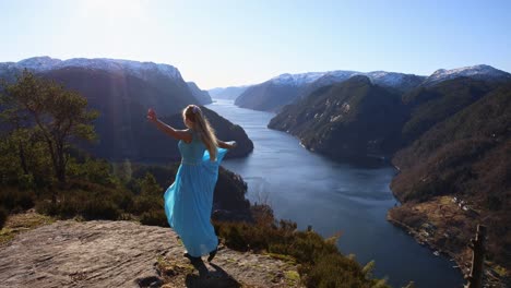 young attractive woman spins, dances on bluff overlooking norwegian fjord in flowing light blue dress on sunny morning.