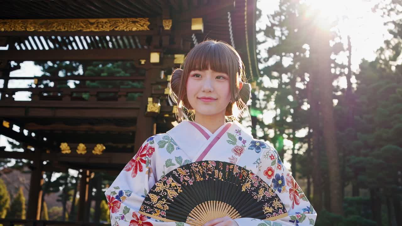 A video still of a woman in a floral kimono holding a fan, captured from a low angle with sunlight
