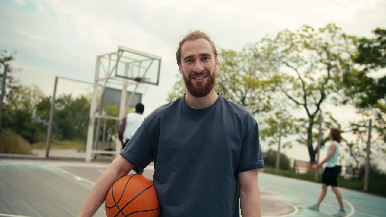 toma de primer plano de un hombre pelirrojo en una camiseta gris con una pelota de baloncesto en las manos posando contra el fondo de un equipo que juega baloncestoen la cancha de baloncestro afuera