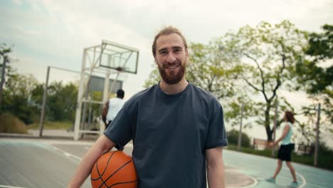 toma de primer plano de un hombre pelirrojo en una camiseta gris con una pelota de baloncesto en las manos posando contra el fondo de un equipo que juega baloncestoen la cancha de baloncestro afuera