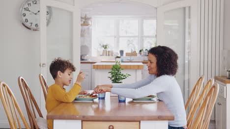 Madre-Soltera-Ayudando-A-Su-Hijo-A-Cortar-La-Comida-Mientras-Se-Sientan-A-La-Mesa-Comiendo-En-Casa