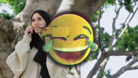 animation of happy emoji over woman in hijab using smartphone