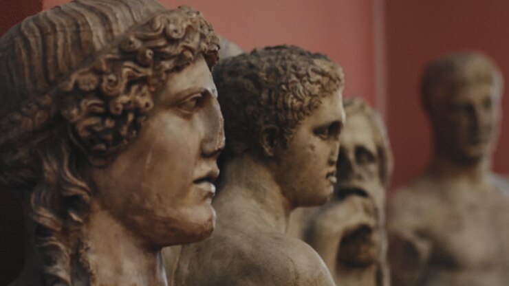 Primo piano di teste di più antiche statue maschili in un museo con luci soffuse e calde.