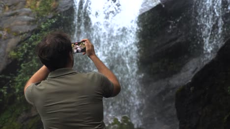 travel man with mobile phone shooting video of waterfall in dark rainforest