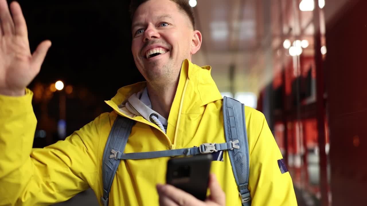 Hombre feliz con smartphone sonríe y saluda a amigos con la mano levantada al aire libre
