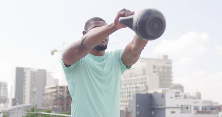 Mid-adult African man starting seated on rooftop railing, swinging kettlebell in reps for strength