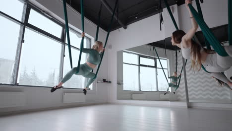 trainer and young woman practising aero gymnastics with suspensions at gym. group of two young beautiful yogi women doing aerial yoga practice in green hammocks in fitness club