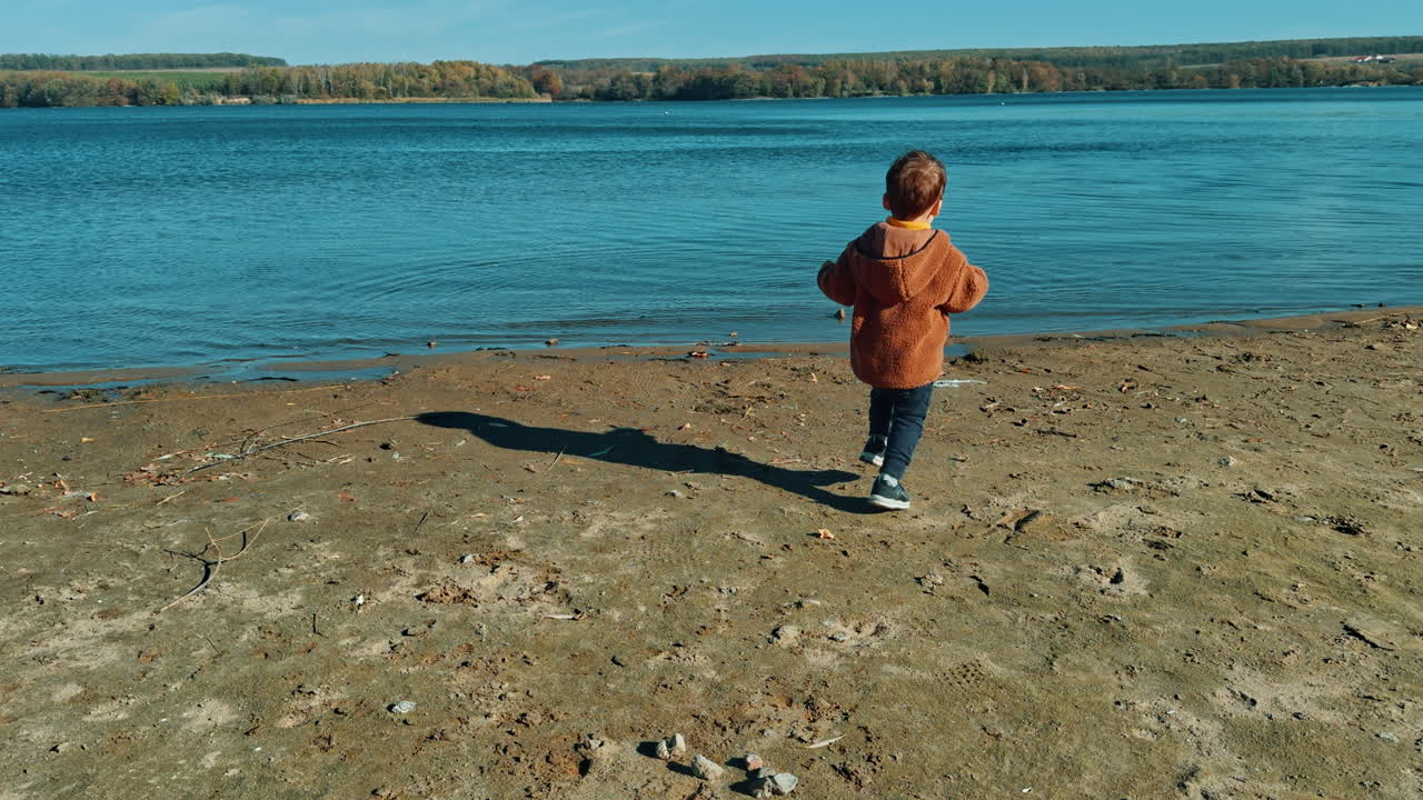 Active Caucasian toddler boy spending time at the river bank on sunny autumn day. Kid picks up stone and throws it into water.
