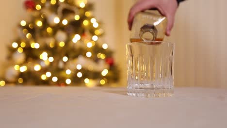 verter whisky a mano en un vaso de cristal en una mesa de cocina celebrando frente a un árbol de navidad iluminado y decorado en casa, cámara lenta