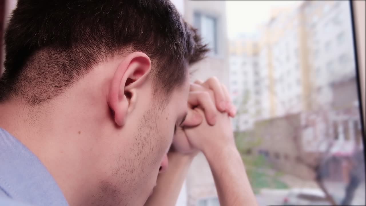 Bored man in blue shirt looking desperate out of the window while keeping hand on the cheek. Close up. Quarantine and Corona Virus idea