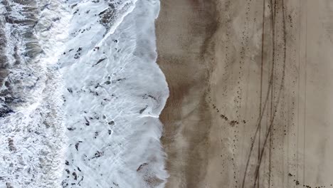 cenital spiral drone shot of the sea waves breaking on the beach with tracks on the sand, in cariló, argentina