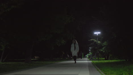 young lady wearing white coat strolls alone in quiet park at night, subtle presence trailing behind her under soft lamp glow, capturing calm mood yet hint of tension, trees shadowy