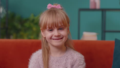 Portrait-of-little-adorable-child-girl-sitting-alone-on-sofa-at-home-looking-at-camera-and-smiling