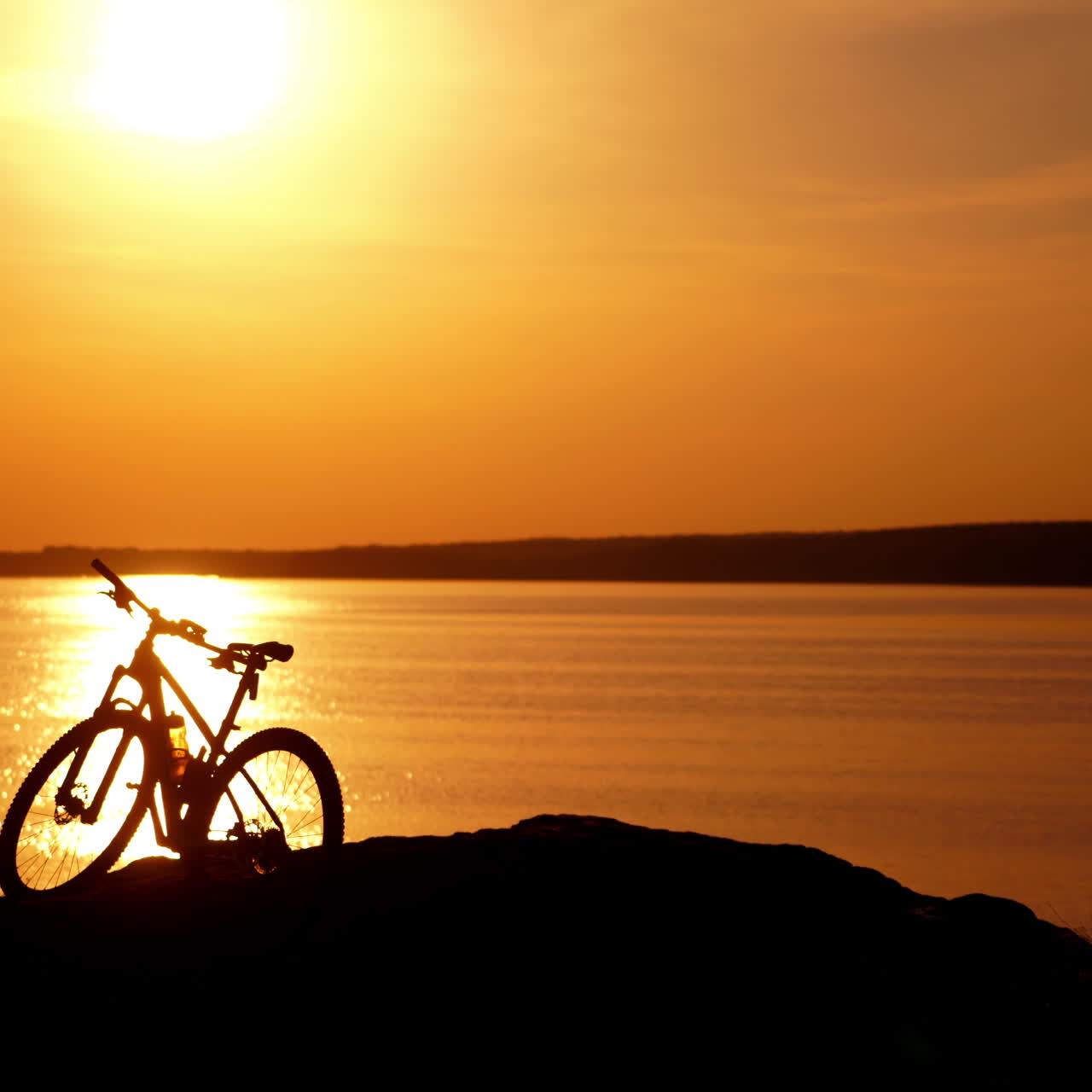 Bicycle and professional cyclist at sunset landscape. Silhouette of a man in helmet comes to the river next to his bike in the evening. Evening orange sun reflects on the water surface.