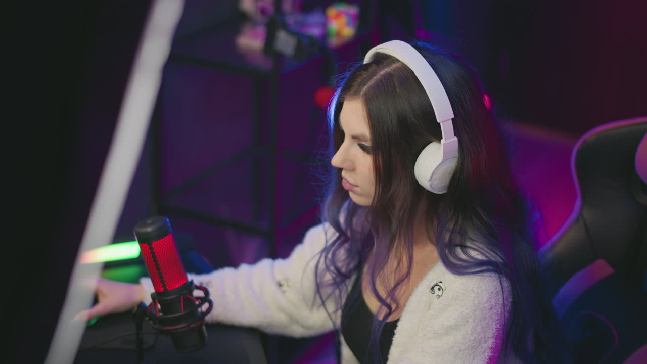 focused young woman with long hair wearing white headphones uses computer mouse in vibrant gaming room, seated in gaming chair with condenser mic in front and neon-lit background full of equipment