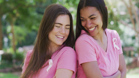 two happy diverse women in pink t shirts and cancer ribbons, talking and laughing in park