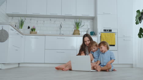 madre apartamento moderno y dos hijos sentados en el suelo en la sala de estar miran la pantalla del portátil