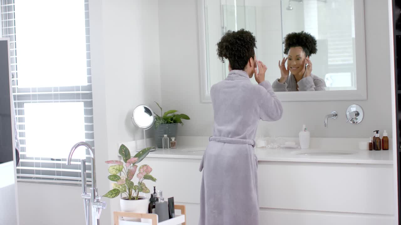 Une femme métisse heureuse en peignoir se regardant dans le miroir dans la salle de bain, au ralenti