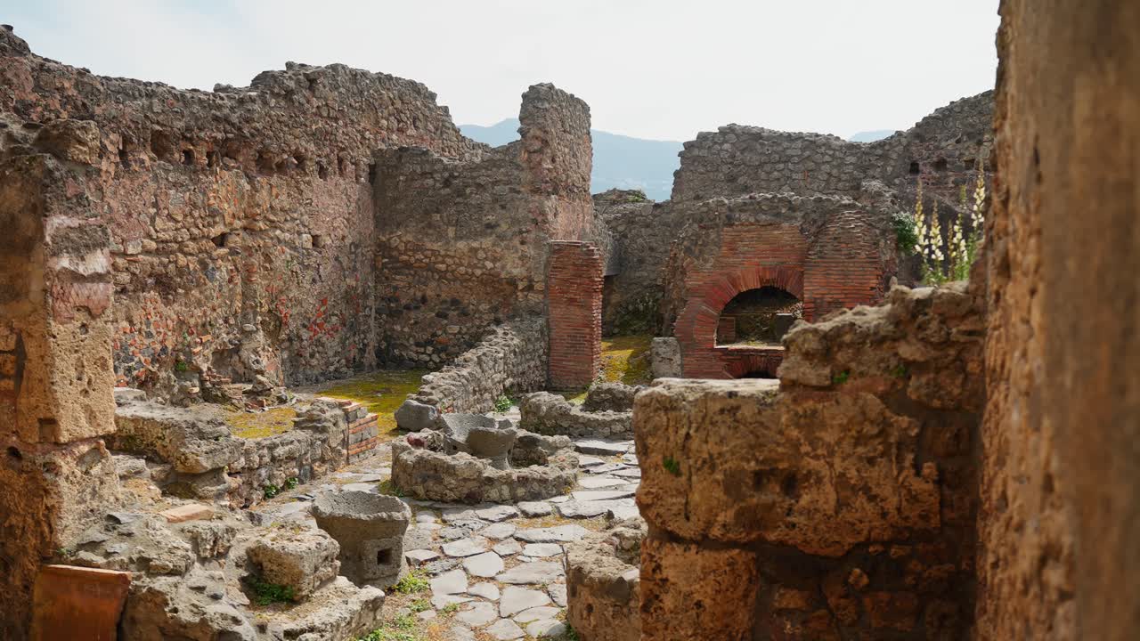 Vista incorniciata delle rovine di Pompei con un forno in mattoni, muri in pietra crollati e muratura romana scavata, che rivela un antico cortile domestico o di officina