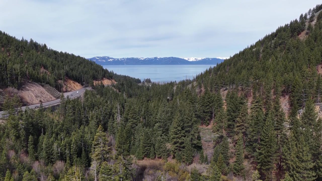 Splendida vista panoramica del Lago Tahoe attraverso una fitta valle di pini, lago alpino di un blu intenso, montagne della Sierra Nevada innevate e strada di montagna tortuosa sotto un cielo grigio e coperto. Ripresa aerea in ascesa