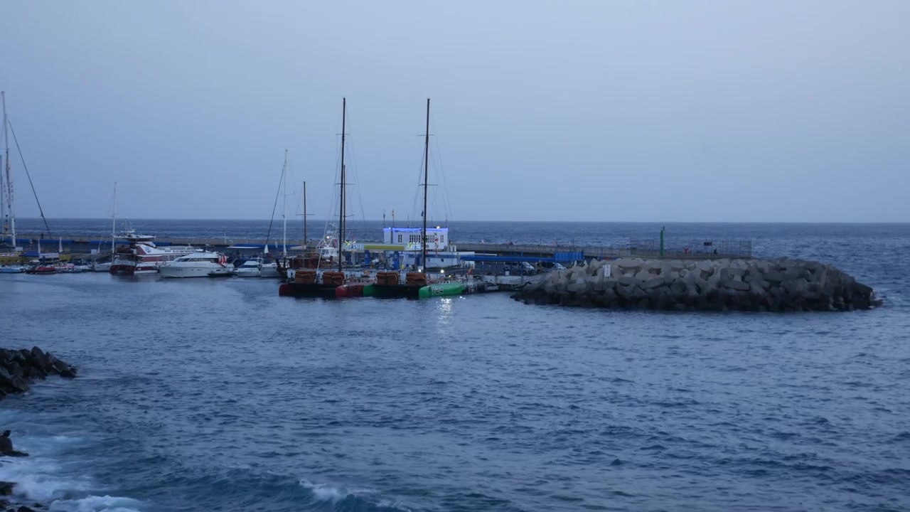 Barche e barche a vela ormeggiate in un porto turistico accanto a un frangiflutti roccioso a Costa Adeje, Tenerife. La vista costiera nell'ora blu mostra le acque calme del porto che incontrano l'Oceano Atlantico