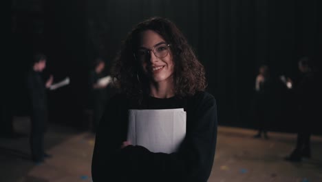 Portrait-of-a-confident-brunette-girl-with-curly-hair-in-glasses-and-a-black-actors-suit-with-papers-in-her-hands-in-a-group-of-actors-who-are-rehearsing-before-performing-on-stage-in-a-theater-with-black-curtains