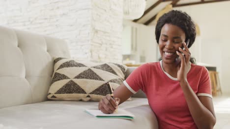 video of happy african american woman sitting on floor, using smartphone and making notes