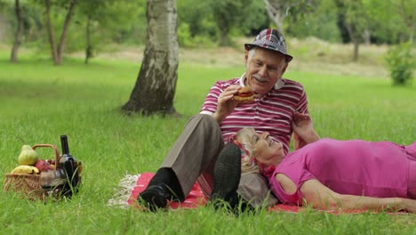 Family-weekend-picnic-in-park.-Active-senior-old-caucasian-couple-sit-on-blanket-and-eating-burger